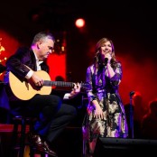 Keith and Kristyn Getty sitting down on chairs on a Stage performing. Keith holds an acoustic guitar and Kristyn is holding a Microphone.